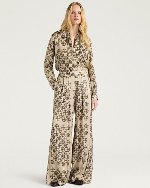 Front view of a cream-colored matching set featuring a long-sleeve button-front shirt and wide-leg trousers, both decorated with an allover black geometric floral-inspired pattern. The shirt has an open collar and a relaxed fit, while the trousers feature a high waist and fluid, wide-leg silhouette. Shown on a model against a light gray background.