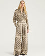 Front view of a cream-colored matching set featuring a long-sleeve button-front shirt and wide-leg trousers, both decorated with an allover black geometric floral-inspired pattern. The shirt has an open collar and a relaxed fit, while the trousers feature a high waist and fluid, wide-leg silhouette. Shown on a model against a light gray background.