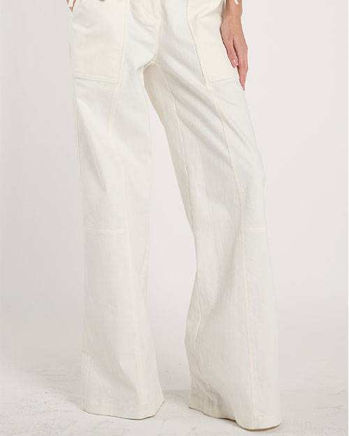 Person standing in a studio against a light neutral background, wearing a coral off-the-shoulder top with a wide ruffled neckline paired with high-waisted white wide-leg pants.