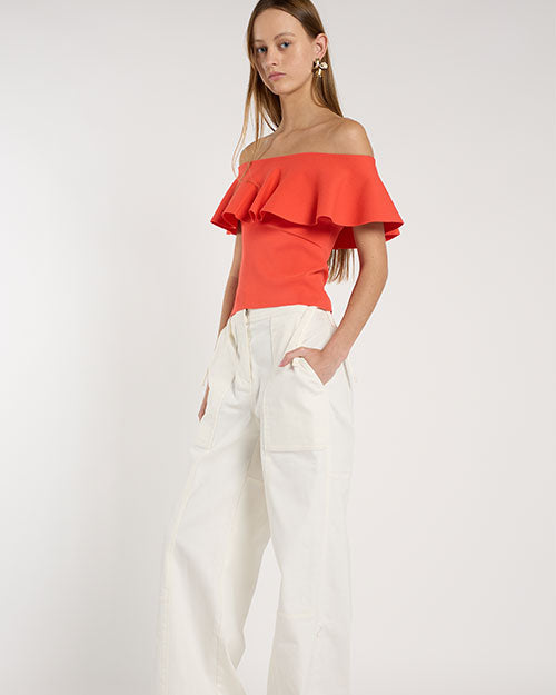 Person standing in a studio against a light neutral background, wearing a coral off-the-shoulder top with a wide ruffled neckline paired with high-waisted white wide-leg pants.