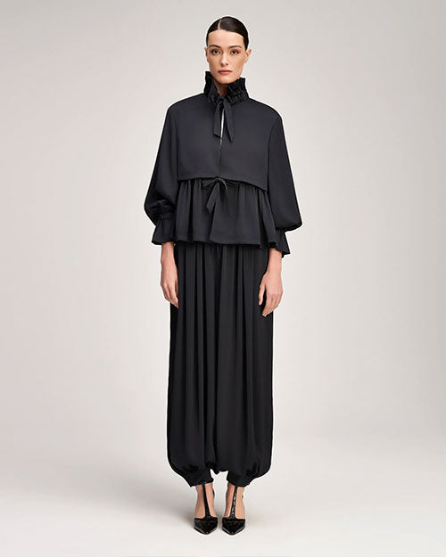 Person wearing a black ruffle-neck peplum top with a tie-front detail, voluminous sleeves, and matching wide-leg pants with gathered hems, paired with pointed black shoes.