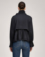 Back view of a black peplum jacket with a ruffled high collar, gathered back panel, and long sleeves with ruffle cuffs, paired with blue jeans.