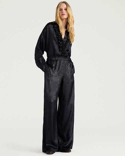 Full-length studio image of a person wearing a black satin-like outfit consisting of a ruffled V-neck blouse and matching wide-leg pants. The long-sleeve top features cascading ruffle detailing along the neckline and front placket, while the loose-fitting pants have an elasticized waistband and a relaxed silhouette. The person stands against a light gray background with one hand in a pocket.