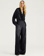 Full-length studio image of a person wearing a black satin-like outfit consisting of a ruffled V-neck blouse and matching wide-leg pants. The long-sleeve top features cascading ruffle detailing along the neckline and front placket, while the loose-fitting pants have an elasticized waistband and a relaxed silhouette. The person stands against a light gray background with one hand in a pocket.