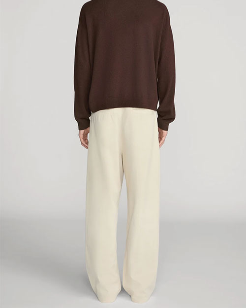 Back view of cream-colored wide-leg trousers with a relaxed fit and full-length legs that fall loosely to the floor. The trousers are paired with a dark brown long-sleeve knit top and shown from the shoulders to the feet against a light gray background.