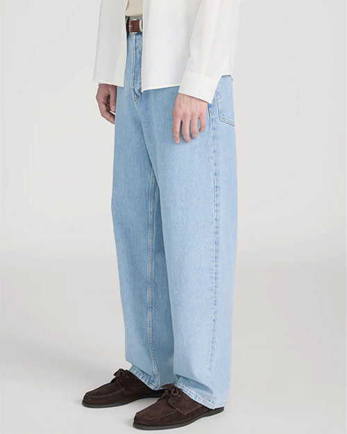 Close-up studio image against a light neutral background, showing light-wash wide-leg jeans with visible seam detailing down the front of each leg. The jeans are styled with a brown leather belt and paired with dark brown suede lace-up shoes. The hem falls loosely over the shoes, and the image is photographed from waist to feet in a slightly angled view.