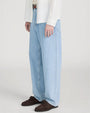 Close-up studio image against a light neutral background, showing light-wash wide-leg jeans with visible seam detailing down the front of each leg. The jeans are styled with a brown leather belt and paired with dark brown suede lace-up shoes. The hem falls loosely over the shoes, and the image is photographed from waist to feet in a slightly angled view.