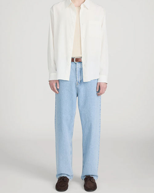 Person standing in a studio against a light neutral background, wearing a light beige crewneck T-shirt layered under an off-white button-down shirt worn open. The outfit is paired with light-wash straight-leg jeans, a brown leather belt, and dark brown suede shoes, photographed in a full-length, front-facing pose.