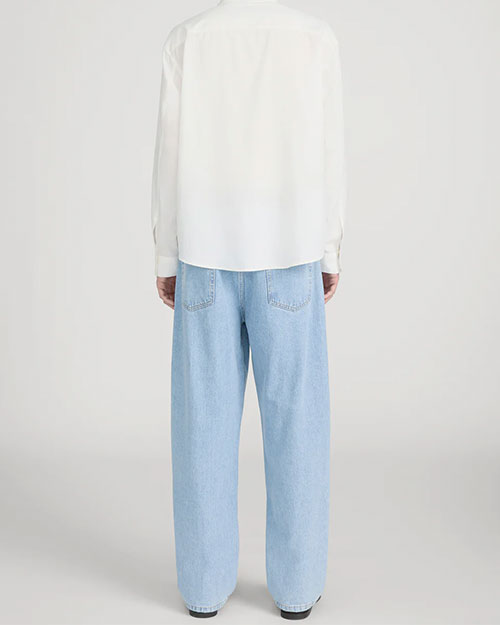 Back view of a person standing in a studio against a light neutral background, wearing a light beige crewneck T-shirt layered under an off-white button-down shirt worn open. The outfit is paired with light-wash straight-leg jeans, a brown leather belt, and dark brown suede shoes, photographed in a full-length, front-facing pose.