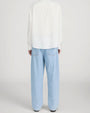 Back view of a person standing in a studio against a light neutral background, wearing a light beige crewneck T-shirt layered under an off-white button-down shirt worn open. The outfit is paired with light-wash straight-leg jeans, a brown leather belt, and dark brown suede shoes, photographed in a full-length, front-facing pose.