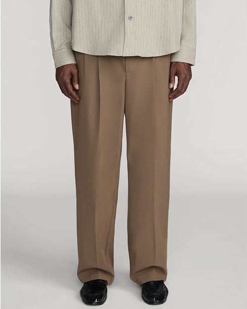 Front view of taupe straight-leg trousers with a relaxed fit and pressed front creases. The trousers are shown from the waist to the feet and are styled with a light beige textured button-front shirt and black leather shoes against a light gray background.