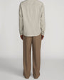 Back view of taupe straight-leg trousers with a relaxed fit and pressed leg creases. The trousers are paired with a light beige textured long-sleeve shirt featuring a subtle vertical stripe pattern and button cuffs. Shown from the shoulders to the feet against a light gray background.