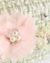 Close-up of a light beige tweed handbag decorated with a pink fabric flower featuring pearl accents, a cream flower, and a pearl embellishment with the word ‘LOVE’ printed on it.