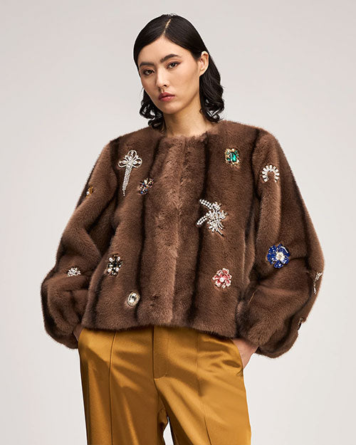 Brown fur-textured jacket embellished with jeweled floral and starburst appliqués, featuring a collarless neckline and voluminous sleeves, paired with satin gold trousers.