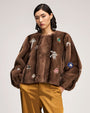 Brown fur-textured jacket embellished with jeweled floral and starburst appliqués, featuring a collarless neckline and voluminous sleeves, paired with satin gold trousers.