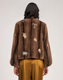 Brown fur-textured jacket embellished with jeweled floral and starburst appliqués, featuring a collarless neckline and voluminous sleeves, paired with satin gold trousers.