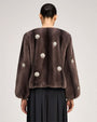 Brown fur-textured jacket with a collarless neckline, voluminous sleeves, and pearl-like floral embellishments throughout, paired with a black gathered skirt.
