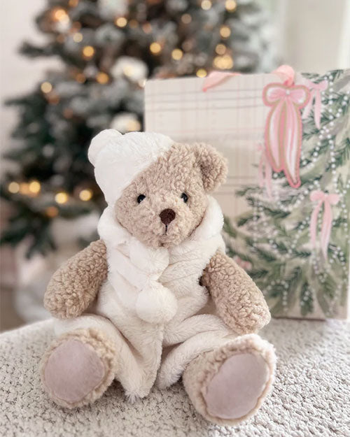  A plush teddy bear dressed in a cozy white winter outfit with fluffy pom-pom accents and a matching hat. The bear has light beige fur, a brown nose, and soft fabric paw pads. It is seated on a textured surface with a blurred Christmas tree and a wrapped gift box featuring a pink ribbon in the background, creating a festive holiday setting.
