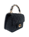 Small black denim handbag with a quilted diamond pattern, gold-tone twist-lock closure, and a short handle featuring interwoven black material and gold chain links, shown in an angled side view.