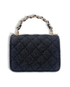 Back view of a small black denim handbag with a quilted diamond pattern and a short handle featuring interwoven black material and gold chain links.