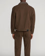 Back view of a model wearing a brown denim-style jacket with a pointed collar, back yoke seam, adjustable button tabs at the hem, and a small black logo patch near the waistband. Paired with matching brown trousers featuring back patch pockets. Shown from the shoulders to the ankles against a light gray background.