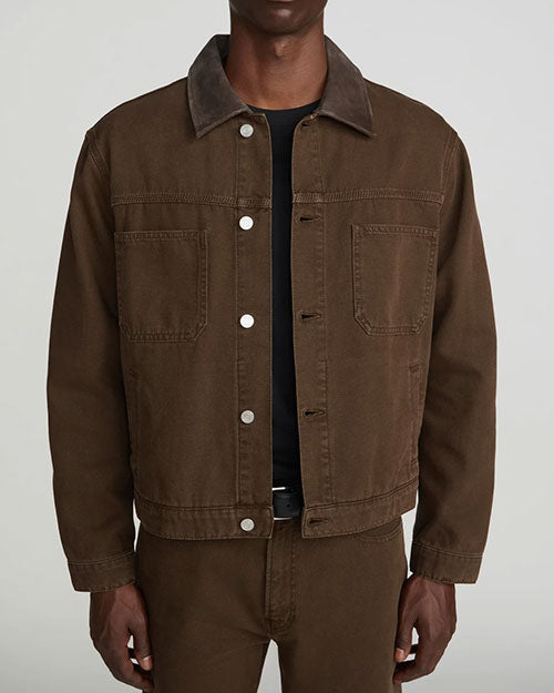 Front view of a model wearing a brown denim-style jacket with a pointed collar, white metal snap-button closure, and two large chest patch pockets. The jacket is layered over a black crewneck T-shirt and paired with matching brown trousers. Shown from the neck to the upper thighs against a light gray background.