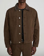 Front view of a model wearing a brown denim-style jacket with a pointed collar, white metal snap-button closure, and two large chest patch pockets. The jacket is layered over a black crewneck T-shirt and paired with matching brown trousers. Shown from the neck to the upper thighs against a light gray background.
