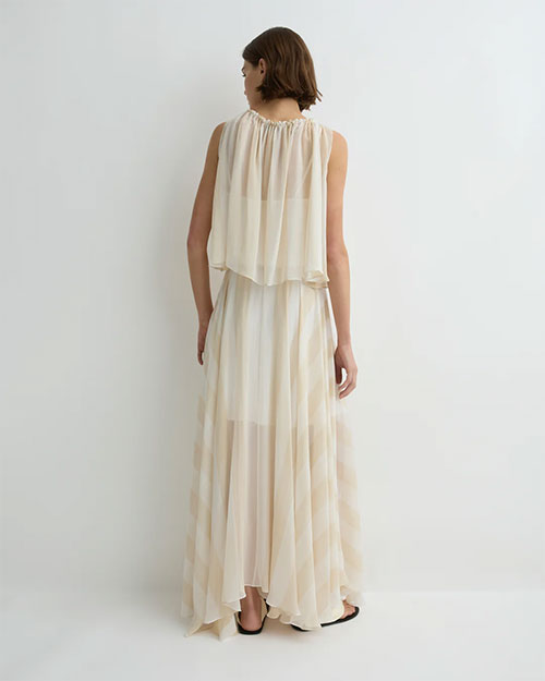 Back view of a person wearing a soft cream sleeveless top with a gathered neckline and layered, flowy fabric, paired with a matching long, airy skirt, against a plain light background.