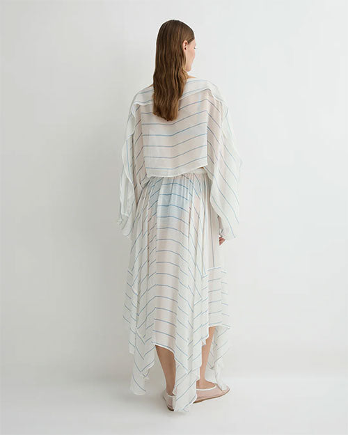 A person standing with their back to the camera, wearing a flowing, sheer long-sleeved top and a matching asymmetrical skirt with light horizontal stripes. The outfit is loose and lightweight, and the person is wearing light-colored flat shoes.