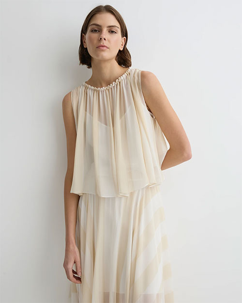 Close-up of a person wearing a soft cream sleeveless top with a gathered neckline and layered, flowy fabric, paired with a matching skirt against a plain light background.