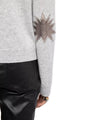 A close-up of a light gray sweater sleeve featuring a dark starburst-shaped elbow patch, shown next to black leather pants.