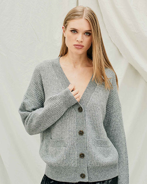 Person wearing a gray textured knit cardigan with a V-neckline and long sleeves. The cardigan features a button-up front with dark buttons and two front pockets. The background is a light fabric backdrop.