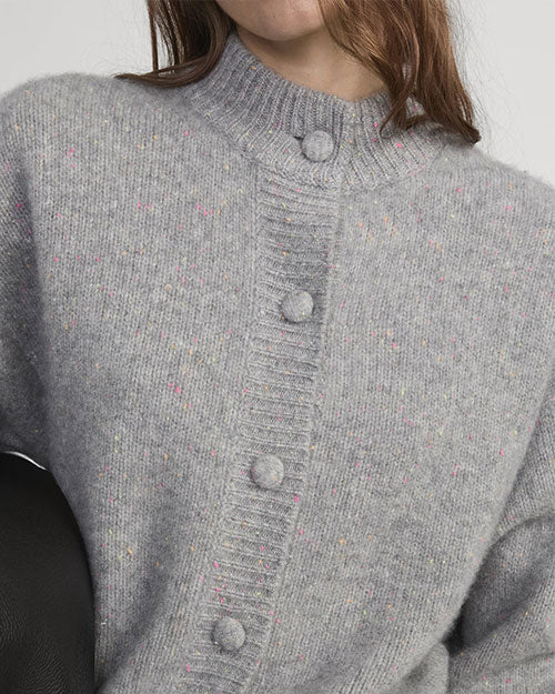 Close-up of a light gray knit cardigan with a ribbed mock neck and fabric-covered buttons, featuring subtle multicolor speckles.