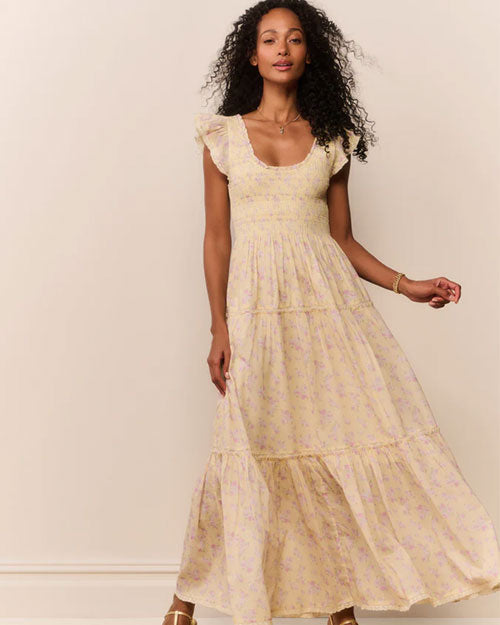 A person wearing a light yellow, ankle‑length dress with a soft floral pattern in pastel tones. The dress features a smocked bodice, a scoop neckline with ruffled edges, and short flutter sleeves. The skirt is tiered with subtle gathered seams, creating a flowing, airy silhouette. The individual is standing against a light-colored background with one arm slightly lifted, causing the skirt to move gently.