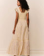 Back view of a person wearing a light yellow, ankle‑length dress with a soft floral pattern in pastel tones. The dress features a smocked bodice, a scoop neckline with ruffled edges, and short flutter sleeves. The skirt is tiered with subtle gathered seams, creating a flowing, airy silhouette. The individual is standing against a light-colored background with one arm slightly lifted, causing the skirt to move gently.