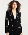 Black tailored blazer adorned with multiple ornate embroidered and jeweled embellishments, including dragonflies, flowers, and decorative motifs in gold, red, and metallic tones. The blazer is worn over a white V-neck top.