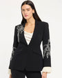 Person wearing a tailored black blazer with white embroidered floral detailing on the lapel and sleeves, layered over a white satin camisole, with matching black trousers, against a light studio background.