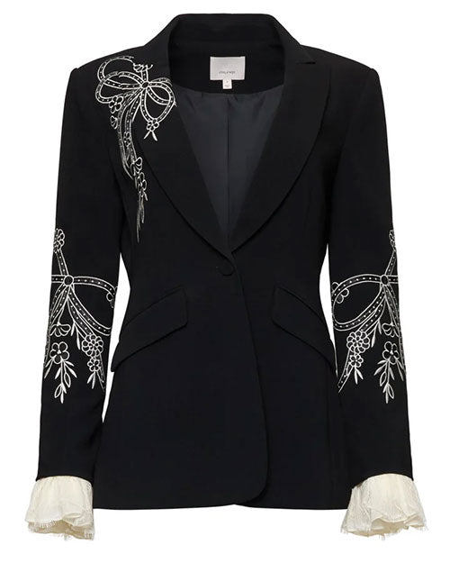 Front view of a tailored black blazer with white embroidered floral and bow detailing on the lapel and sleeves, featuring a single-button closure, contoured seams, flap pockets, and ruffled white cuffs, shown against a white background.