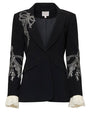 Front view of a tailored black blazer with white embroidered floral and bow detailing on the lapel and sleeves, featuring a single-button closure, contoured seams, flap pockets, and ruffled white cuffs, shown against a white background.