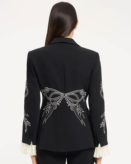 Back view of a tailored black blazer featuring white embroidered bow and floral detailing across the lower back and sleeves, with subtle waist shaping and buttoned cuffs, photographed against a light studio background.