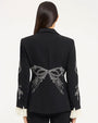 Back view of a tailored black blazer featuring white embroidered bow and floral detailing across the lower back and sleeves, with subtle waist shaping and buttoned cuffs, photographed against a light studio background.