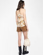 Back view of an outfit featuring a beige camisole top with lace trim and an abstract horse print in shades of brown and tan. The top is paired with high-waisted brown suede shorts and black mid-calf boots. The person is holding a cream-colored drawstring bag with a gold chain strap and decorative tassels.