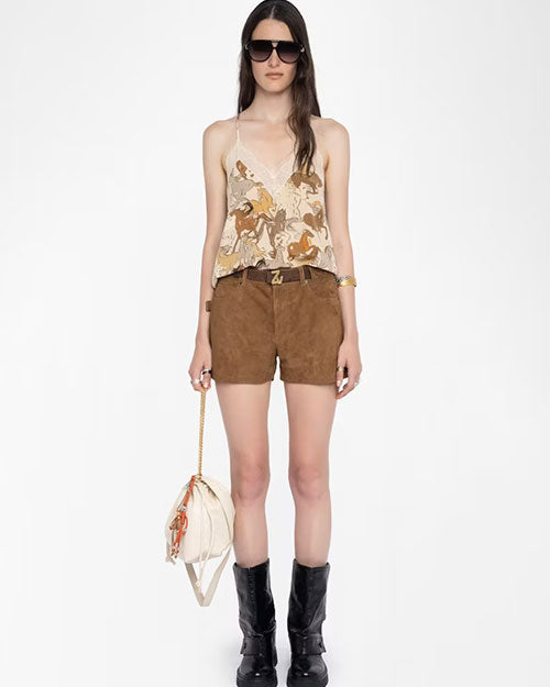 A person wearing a beige camisole top with a lace-trimmed neckline and an abstract horse print in shades of brown and tan. The top is paired with high-waisted brown suede shorts featuring a belt with a gold “Z” buckle. The outfit is styled with black mid-calf boots and accessorized with a cream-colored shoulder bag with a gold chain strap and decorative tassels, along with multiple rings and a bracelet.