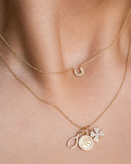 Two gold necklaces worn around a neck. The upper necklace features a small, diamond-encrusted crescent-shaped pendant. The lower necklace includes multiple charms: a circular coin-like charm with an engraved design, an oval-shaped charm encrusted with diamonds, and a diamond-encrusted four-leaf clover charm.