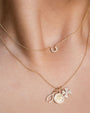 Two gold necklaces worn around a neck. The upper necklace features a small, diamond-encrusted crescent-shaped pendant. The lower necklace includes multiple charms: a circular coin-like charm with an engraved design, an oval-shaped charm encrusted with diamonds, and a diamond-encrusted four-leaf clover charm.