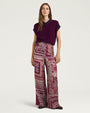 Front view of wide-leg trousers featuring an allover patchwork-inspired print in shades of burgundy, pink, cream, and purple. The high-waisted trousers are styled with a deep plum knit sweater vest with ribbed trim and shown against a light gray background.