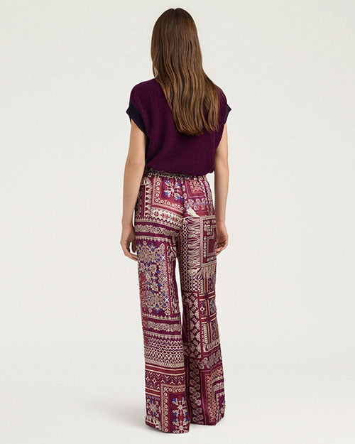 Back view of wide-leg trousers featuring an allover patchwork-inspired print in shades of burgundy, pink, cream, and purple. The high-waisted trousers are styled with a deep plum knit sweater vest with ribbed trim and shown from the shoulders to the feet against a light gray background.