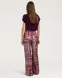 Back view of wide-leg trousers featuring an allover patchwork-inspired print in shades of burgundy, pink, cream, and purple. The high-waisted trousers are styled with a deep plum knit sweater vest with ribbed trim and shown from the shoulders to the feet against a light gray background.