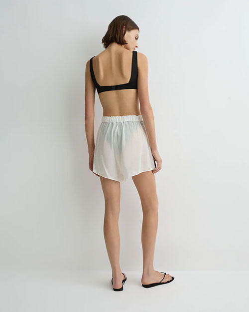 A person standing with their back to the camera, wearing a black sleeveless crop top, loose white shorts with an elastic waistband, and black sandals. They are positioned against a plain light-colored background.