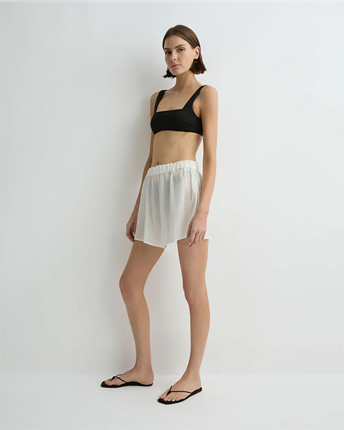 A person  wearing a black sleeveless crop top, loose white shorts with an elastic waistband, and black sandals. They are positioned against a plain light-colored background.